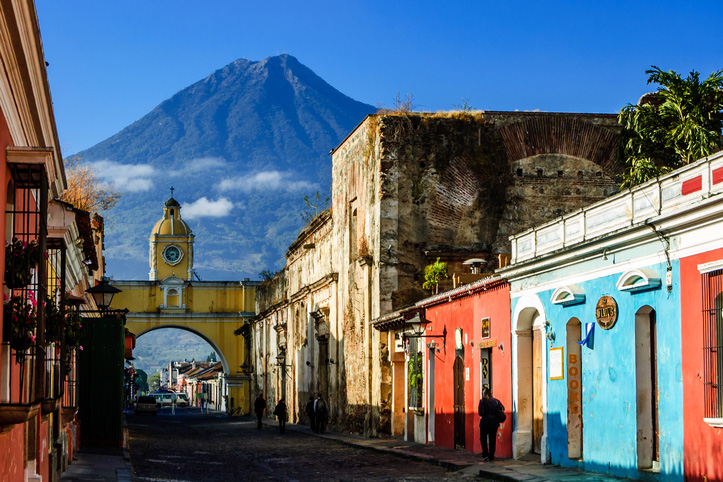 Antigua, Guatemala - March 11, 2012: Locals walk to work along Antigua's famous cobblestoned street lined with painted shops, restaurants & hotels. Agua volcano looms behind Santa Catalina Arch, a landmark in this Spanish colonial town & UNESCO World Heritage Site. Originally the arch allowed nuns to pass from one side of Santa Catalina convent to the other without going outside. Ruins of the convent remain to the right of the arch.